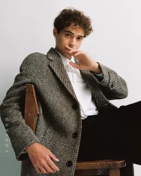 Young man with curly hair wearing a herringbone tweed coat and white shirt seated on a wooden chair against a plain wall