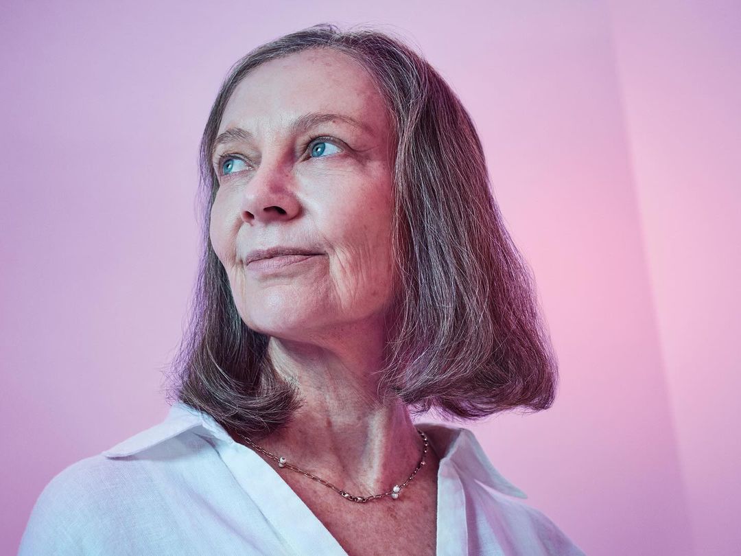 Headshot of an elderly woman with short gray hair wearing a white shirt and necklace against a pink and white gradient background.