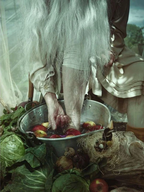 Elderly person washing feet in a basin of apples and vegetables, possibly outdoors. Focus on hands, feet, fruit, and surrounding items.
