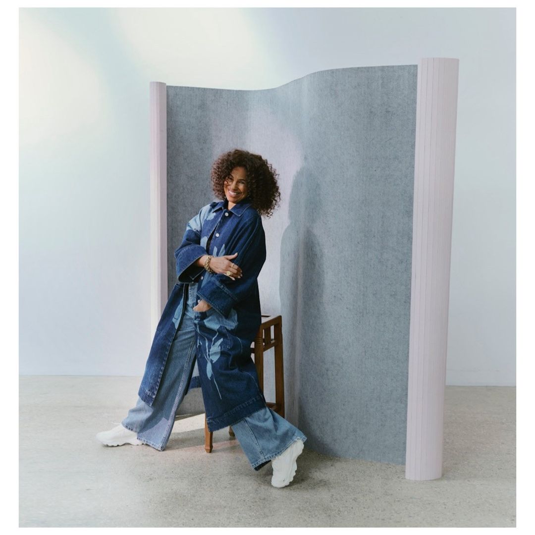 A woman with curly hair is posing indoors, sitting on a stool while wearing a denim coat and denim pants in front of a divider.