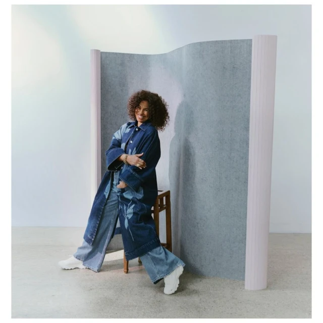 A woman with curly hair is posing indoors, sitting on a stool while wearing a denim coat and denim pants in front of a divider.
