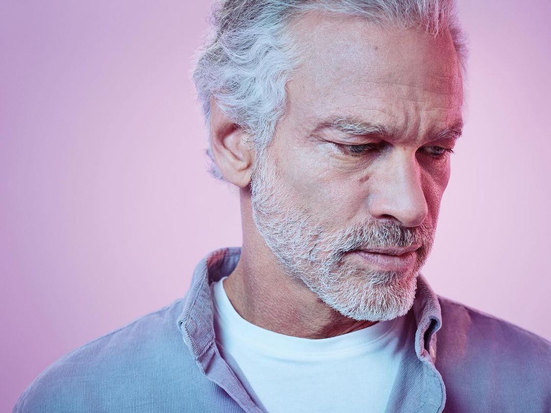 Close-up of a mature adult man with grey hair and beard, wearing a casual shirt, looking down against a soft pink background.