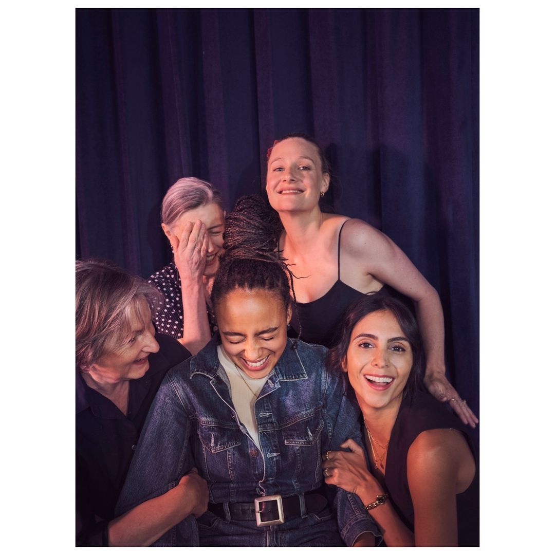 Five women are posing indoors for a group photo. They are smiling and wearing casual clothing. The background is a blue curtain.