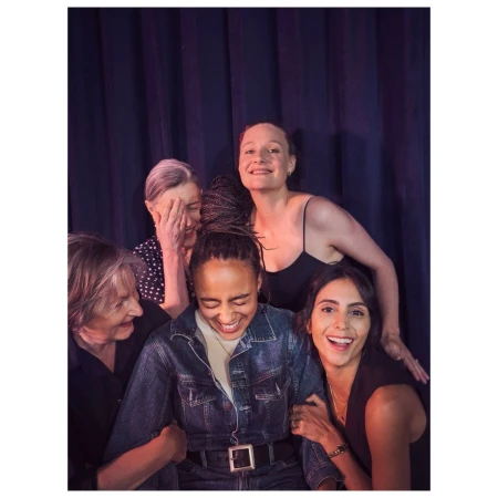 Five women are posing indoors for a group photo. They are smiling and wearing casual clothing. The background is a blue curtain.