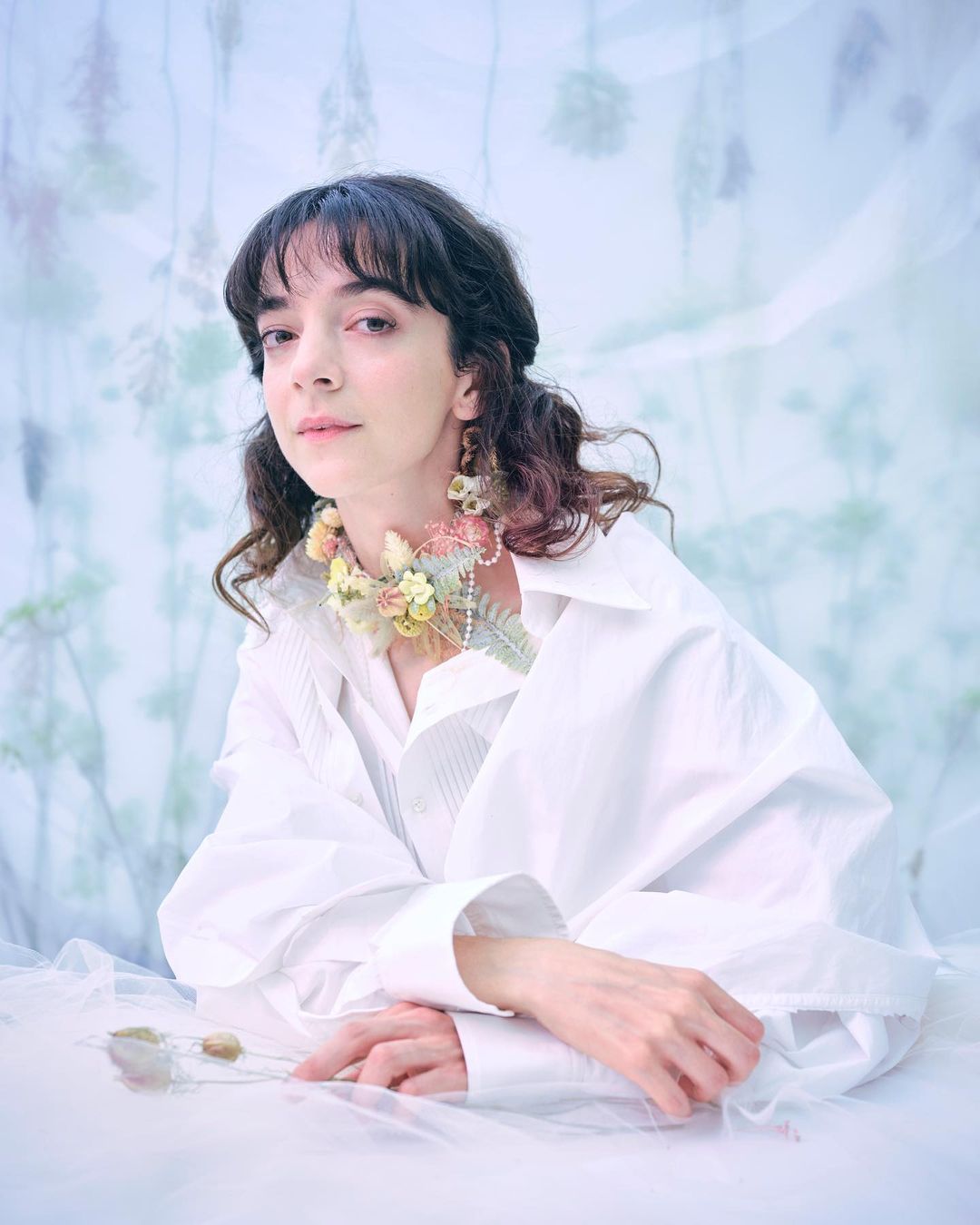 Young woman in white shirt poses with a floral collar and earrings against a blurred, floral backdrop in soft, diffused light.