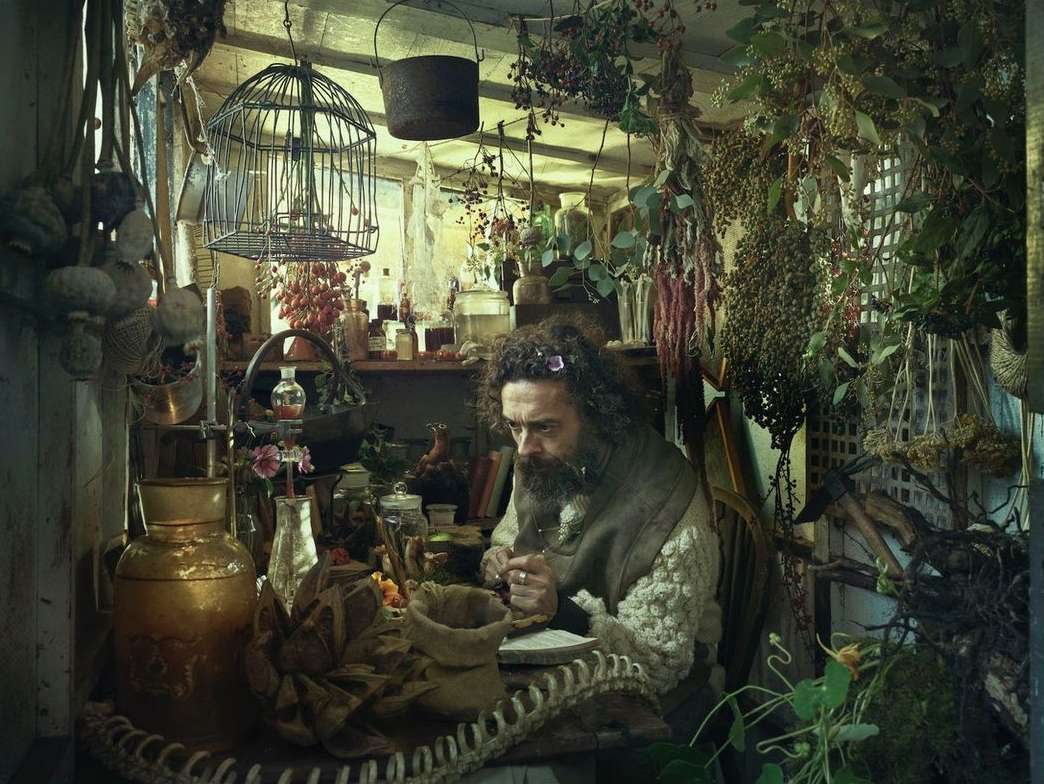 Man with a beard sits at a table filled with jars and herbs inside a rustic room with hanging plants, illuminated by soft, natural lighting.