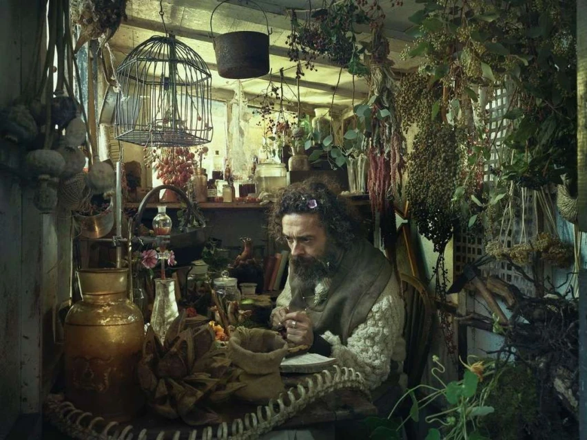 Man with a beard sits at a table filled with jars and herbs inside a rustic room with hanging plants, illuminated by soft, natural lighting.