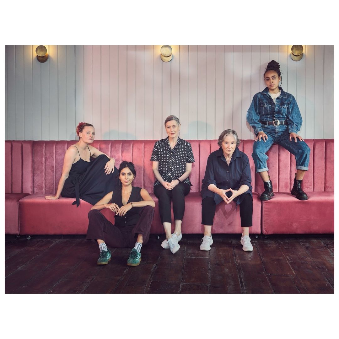Five women pose indoors on a pink velvet couch against a white wall with sconces. They are dressed in casual and formal attire.