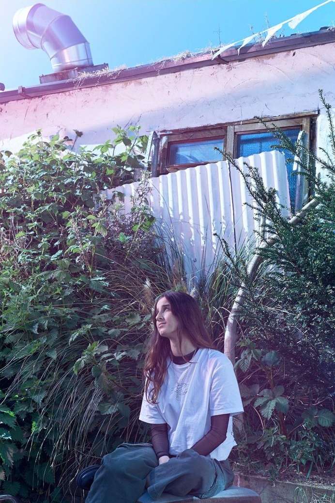 Young woman with long brown hair sits outdoors surrounded by lush greenery and building with a metal vent, bathed in natural light.