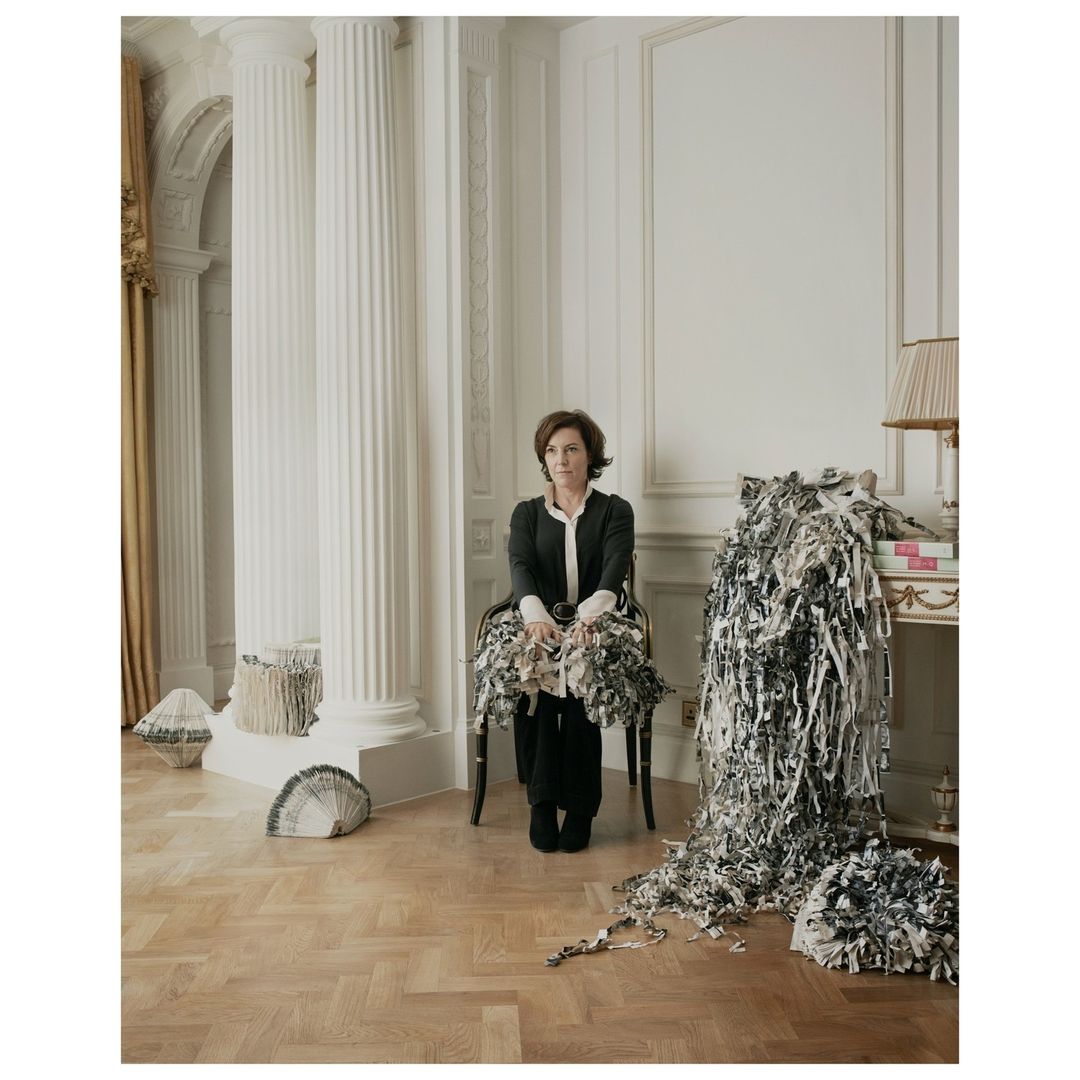 Indoor portrait of a brunette woman seated in a chair amid paper scraps, wearing a black blazer and pants, with a decorative interior backdrop.