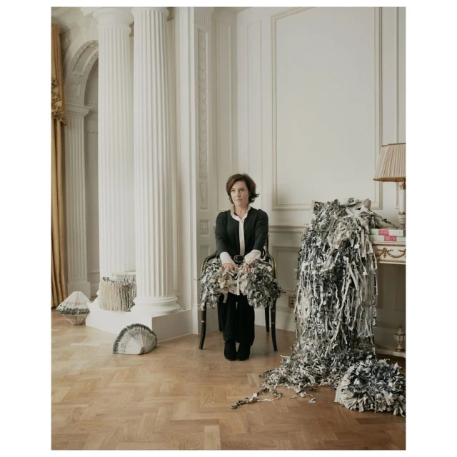 Indoor portrait of a brunette woman seated in a chair amid paper scraps, wearing a black blazer and pants, with a decorative interior backdrop.