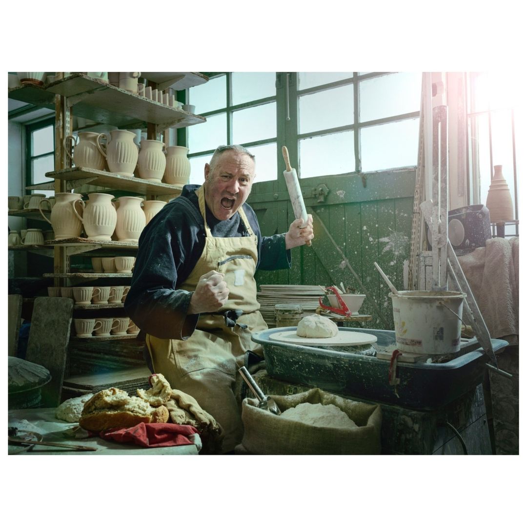 Excited man in pottery studio holding rolling pin. Shelves of pottery surround potter, with visible clay and crafting tools.