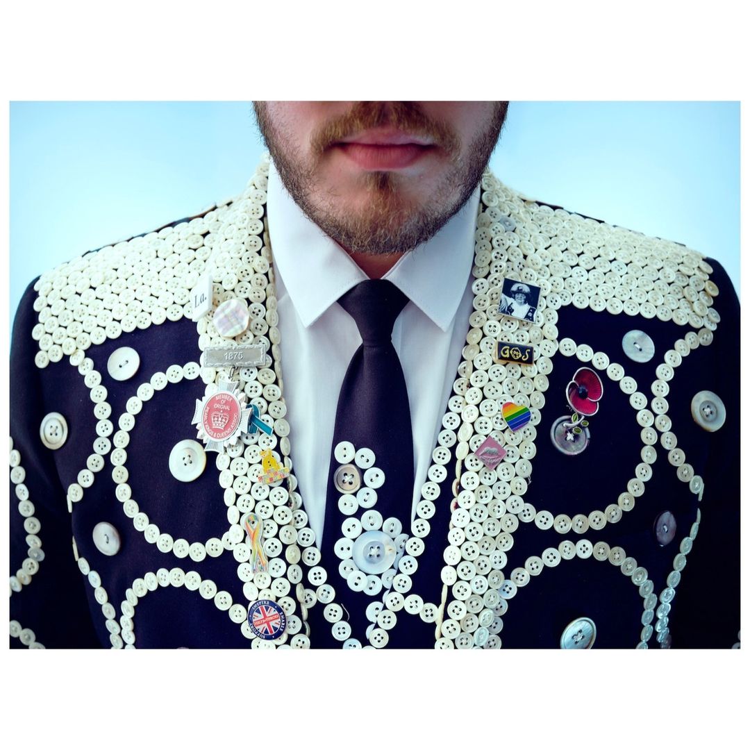 Upper body shot of a man in a white collared shirt and tie, wearing a black jacket adorned with white buttons and pins.