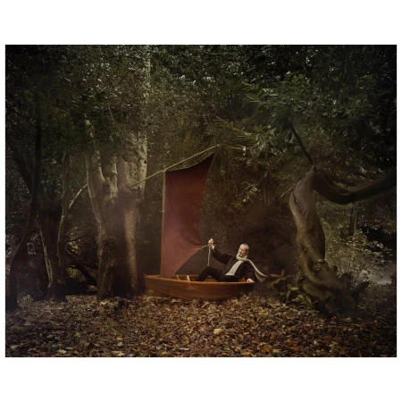 Mature man dressed in suit sitting in a wooden boat with a red sail in the middle of a forest with fallen leaves on the ground.