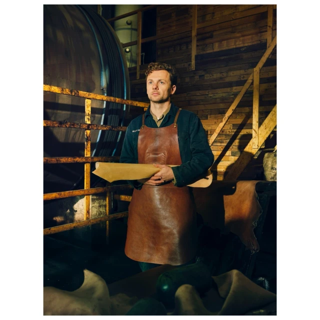 Leather craftsman stands in a workshop wearing a brown apron, holding a piece of leather, with tools and materials visible in the background.