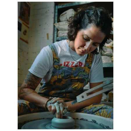Adult female potter works at a pottery wheel, shaping clay, wearing a t-shirt and overalls with a sunflower print in an indoor studio.