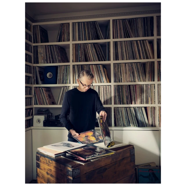 Man in black long-sleeved shirt browses vinyl records in a home interior with shelves of LPs and a wooden chest.