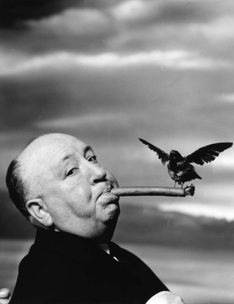 Black and white portrait of a man with a bird perched on a cigar, featuring a cloudy sky background; shot with a medium camera angle.