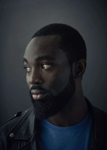 Close-up portrait of a dark-skinned adult man with a beard, wearing a blue shirt and black jacket, looking off to the side in a studio setting.