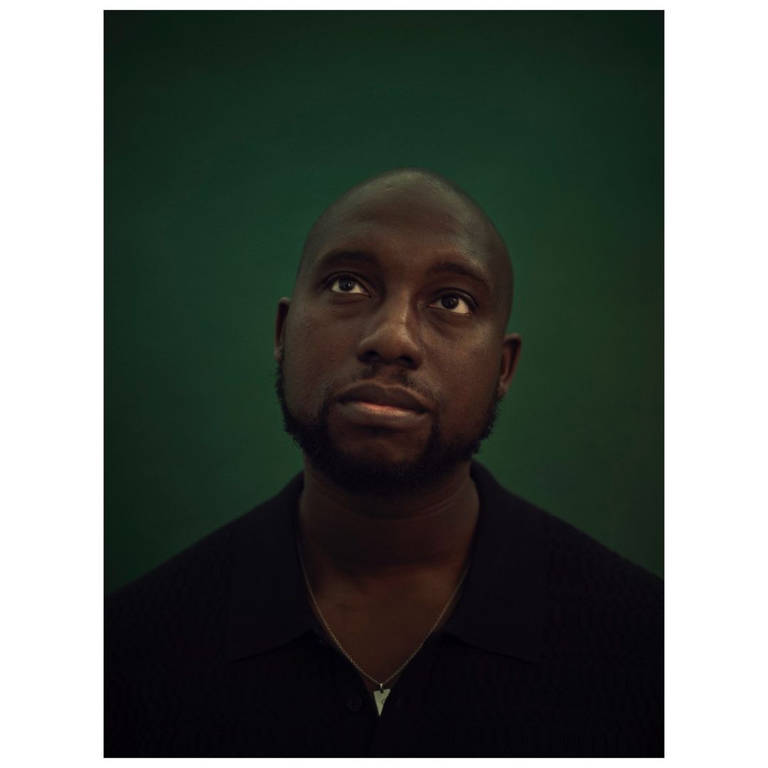 Close-up portrait of a Black man looking up against a green background. He is wearing a black shirt and a silver necklace.
