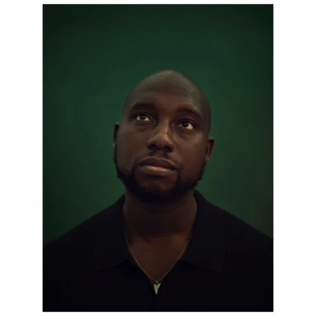 Close-up portrait of a Black man looking up against a green background. He is wearing a black shirt and a silver necklace.