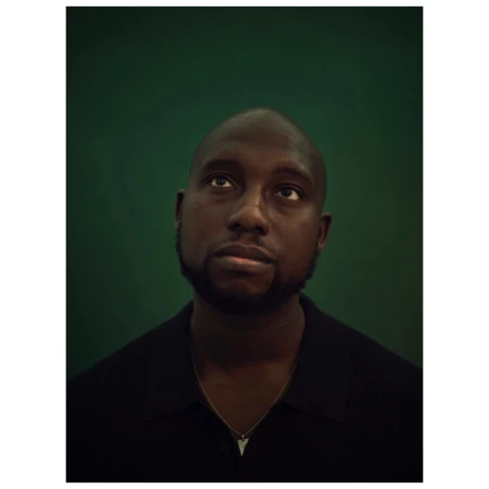 Close-up portrait of a Black man looking up against a green background. He is wearing a black shirt and a silver necklace.