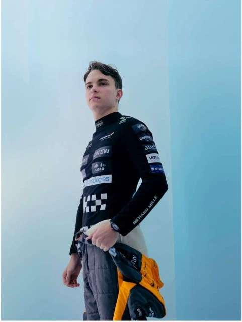 Formula 1 driver stands indoors near gradient wall, wearing a branded black racing suit, holding another suit, medium shot.