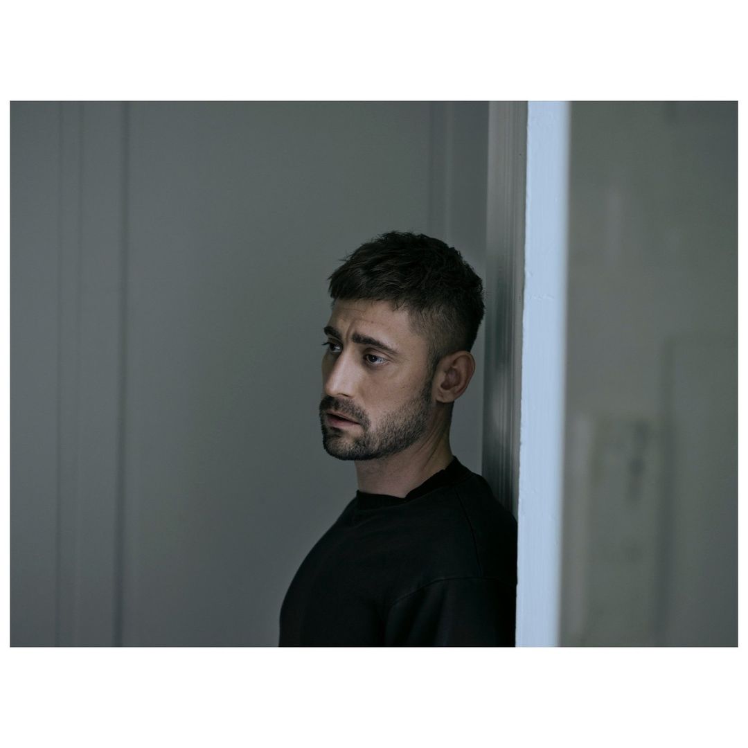 Portrait of an adult male wearing a black shirt, looking away from the camera in an indoor setting with gray walls and soft lighting.