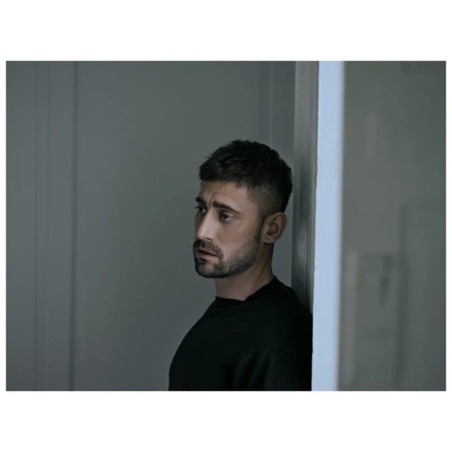 Portrait of an adult male wearing a black shirt, looking away from the camera in an indoor setting with gray walls and soft lighting.