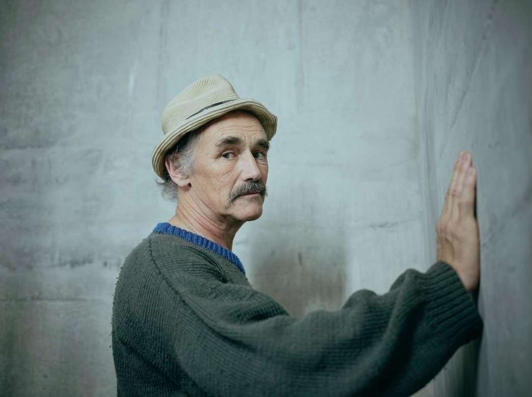 An older adult male with a mustache, wearing a hat and sweater, is standing with his hand against a grey textured wall in a portrait.