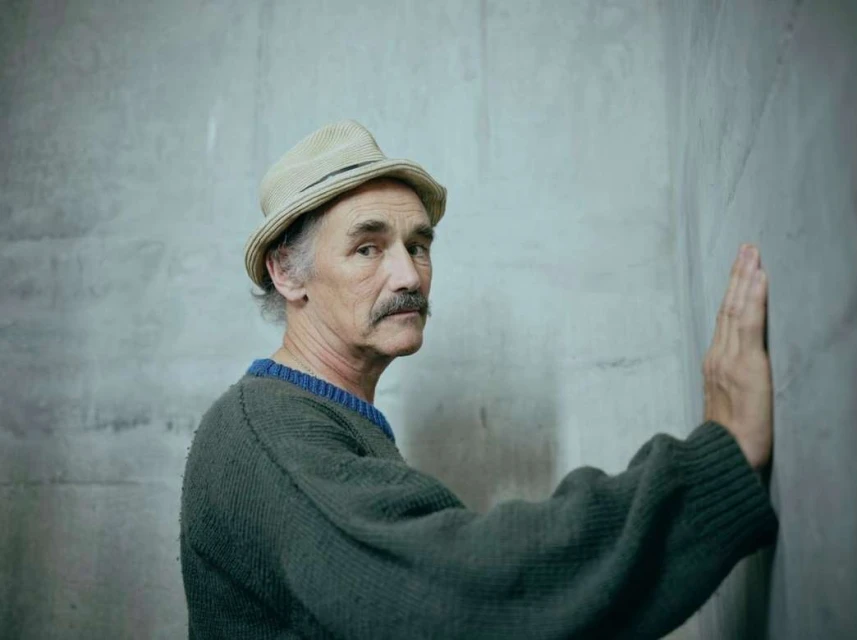An older adult male with a mustache, wearing a hat and sweater, is standing with his hand against a grey textured wall in a portrait.