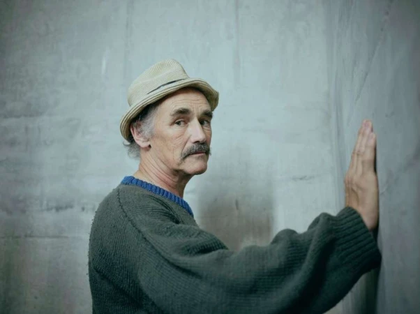 An older adult male with a mustache, wearing a hat and sweater, is standing with his hand against a grey textured wall in a portrait.
