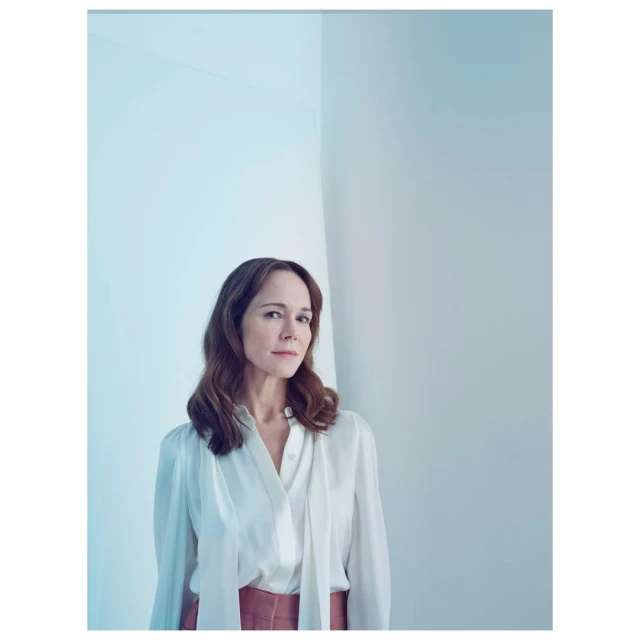Portrait of a woman with long brown hair in a white blouse and pink skirt, looking off to the side in front of a white wall.