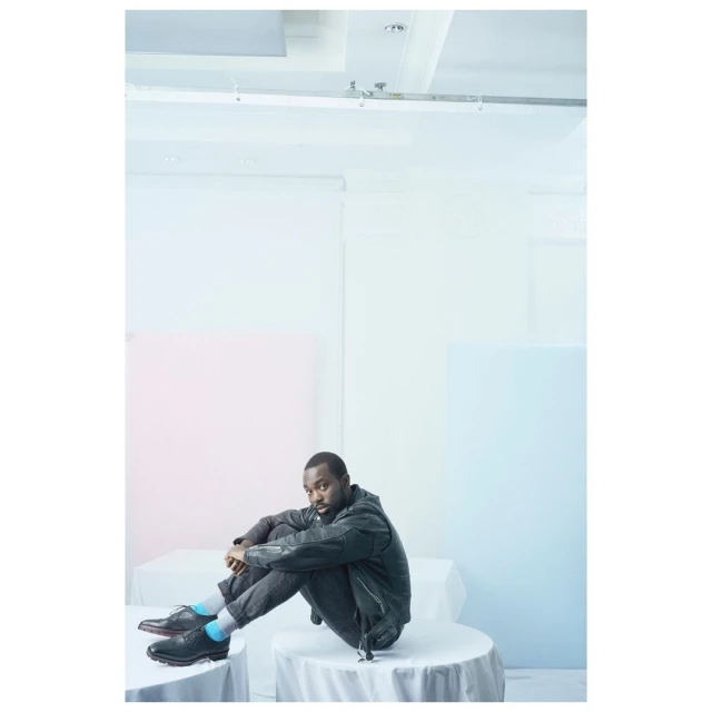 Dark-skinned adult male in black jacket and jeans sits cross-legged on a white-covered round table in a photo studio with colorful pastel backdrops.