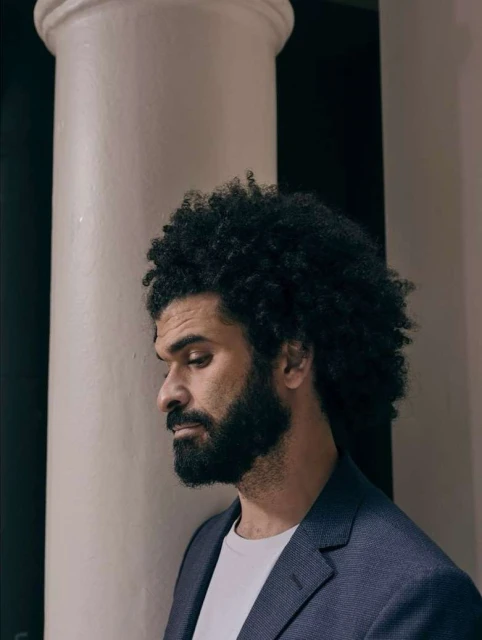 Portrait of a bearded adult man with curly hair in a blue blazer standing near a pillar indoors with soft, neutral lighting.