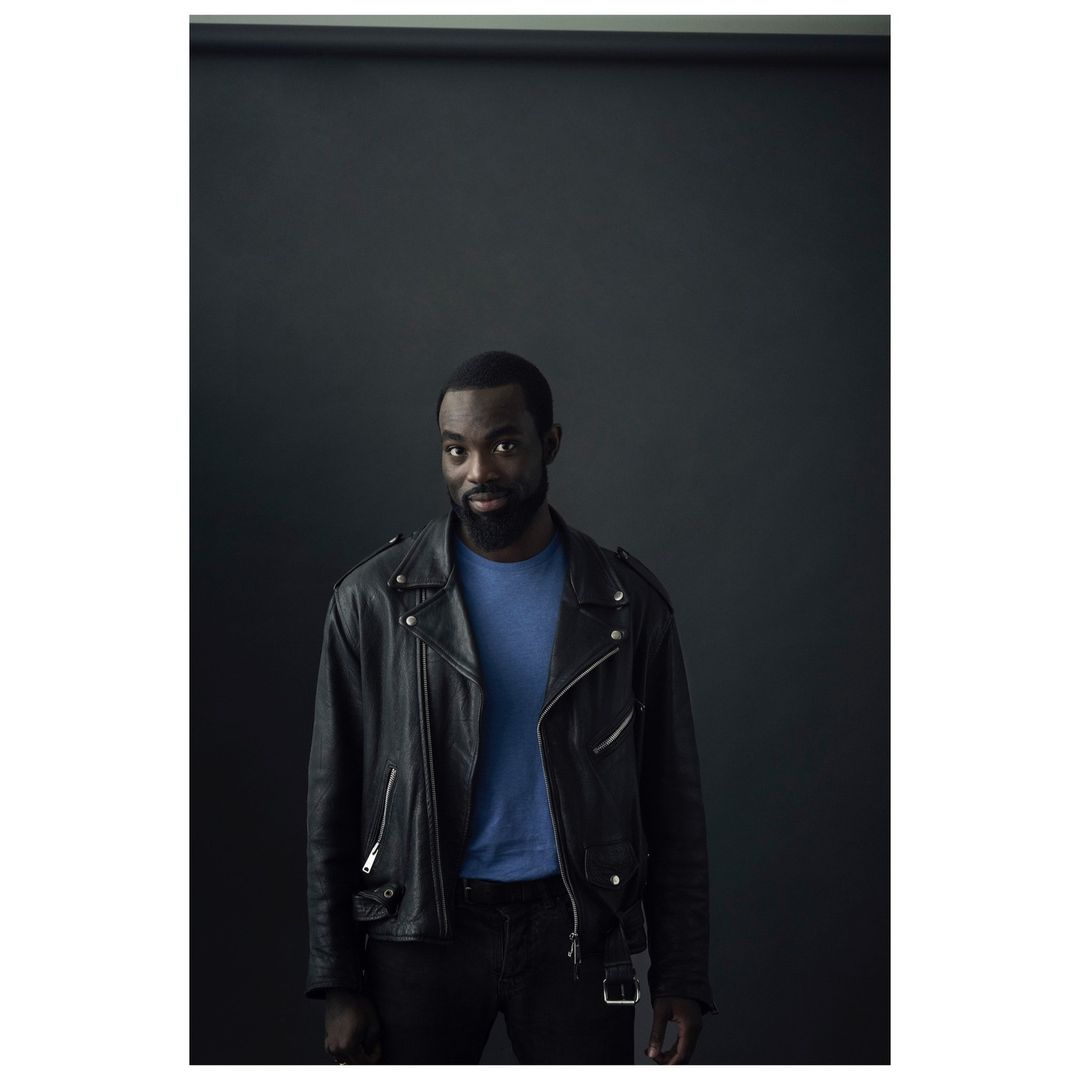 Portrait of a handsome African-American man in a leather jacket. He is wearing a blue shirt and black pants against a dark backdrop.