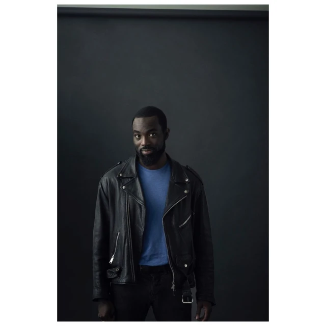 Portrait of a handsome African-American man in a leather jacket. He is wearing a blue shirt and black pants against a dark backdrop.