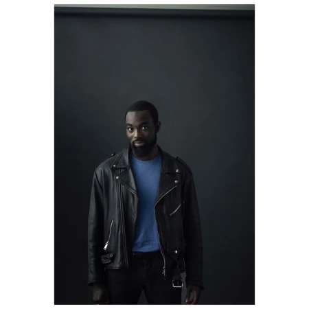 Portrait of a handsome African-American man in a leather jacket. He is wearing a blue shirt and black pants against a dark backdrop.