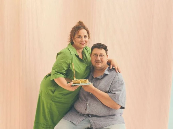 Man and woman holding a small tart on a plate against a pale pink background, photographed by Phil Fisk.