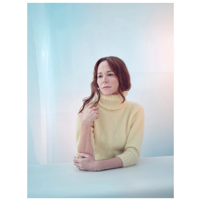 Portrait of a woman with shoulder-length brown hair wearing a yellow turtleneck sweater indoors with soft, diffused lighting. 