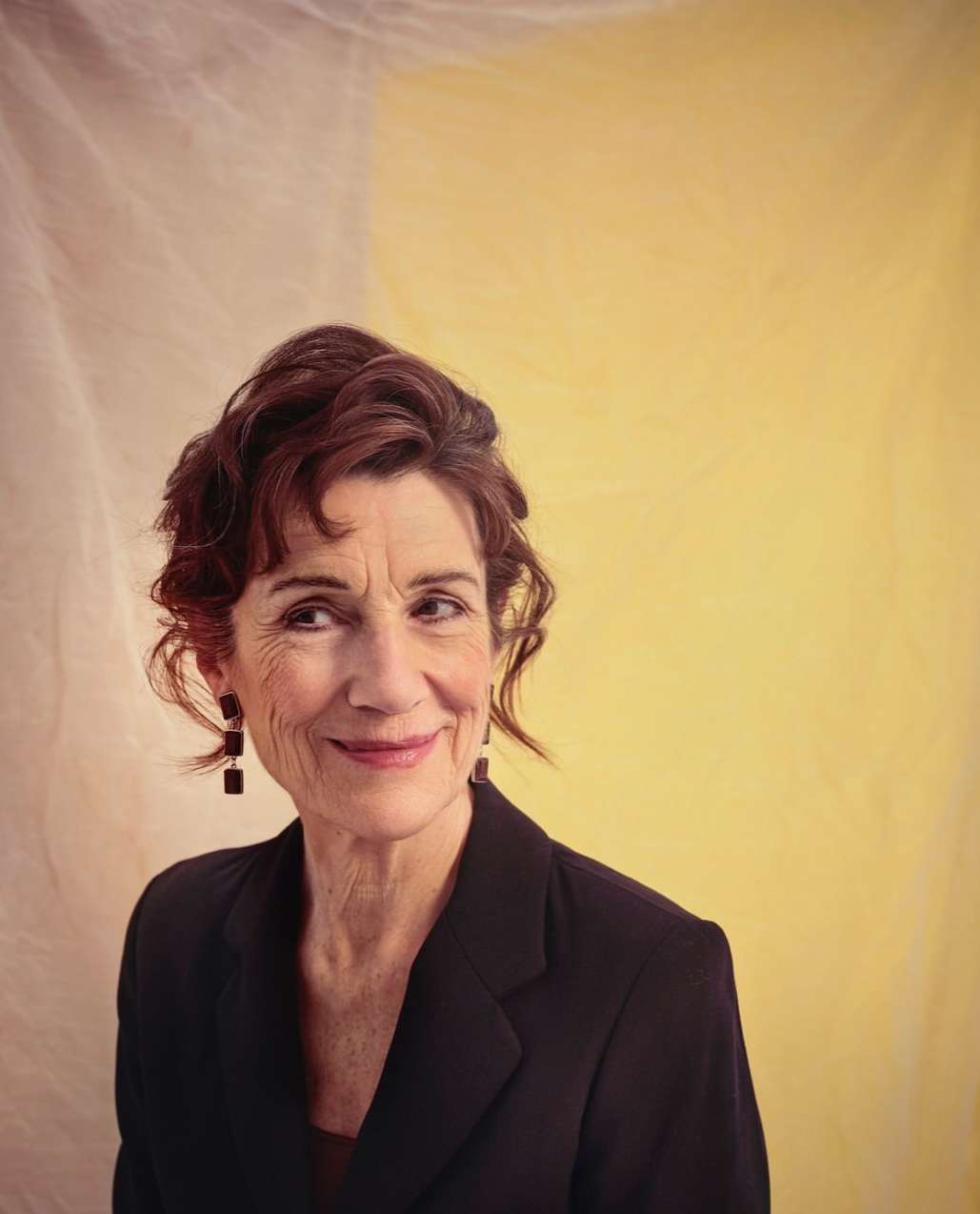 Portrait of a smiling mature woman with auburn hair in a black jacket against a yellow and white mottled background. Head and shoulders.
