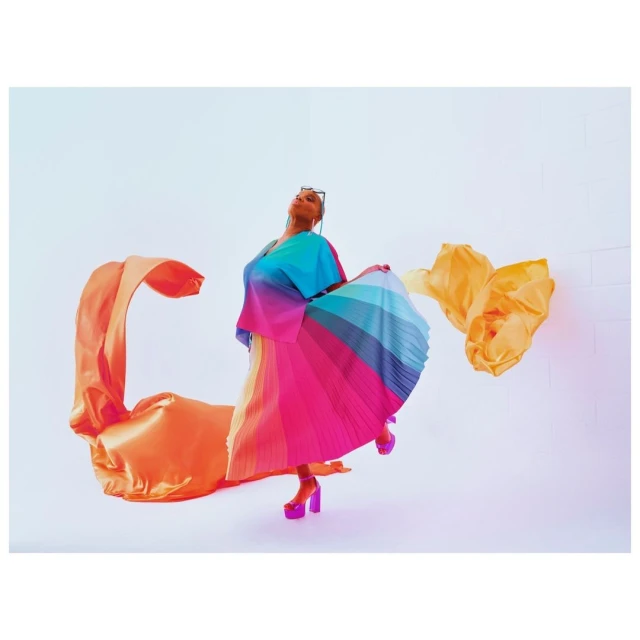 Woman in colorful rainbow dress poses dynamically indoors with flowing fabric. Fashion shot in a bright studio setting.