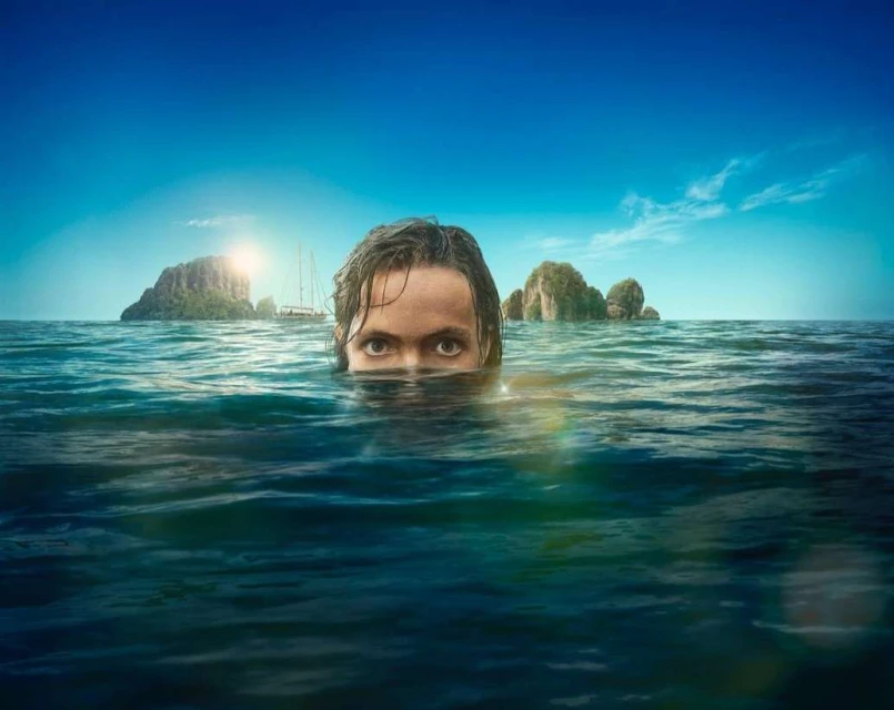 Adult male swimming in ocean water, with islands and a sailboat in the background under a bright blue sky with light cloud cover.
