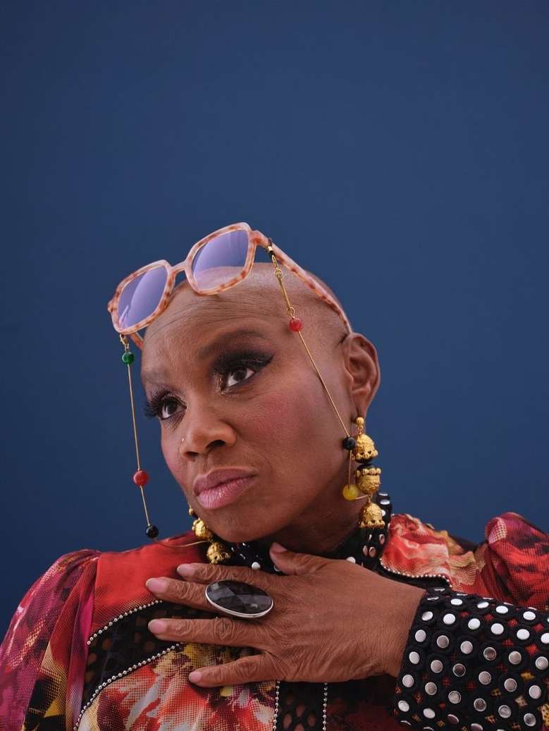 Portrait of a bald Black woman wearing tortoiseshell glasses on her head, ornate earrings, and patterned top against a dark blue background