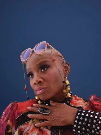 Portrait of a bald Black woman wearing tortoiseshell glasses on her head, ornate earrings, and patterned top against a dark blue background