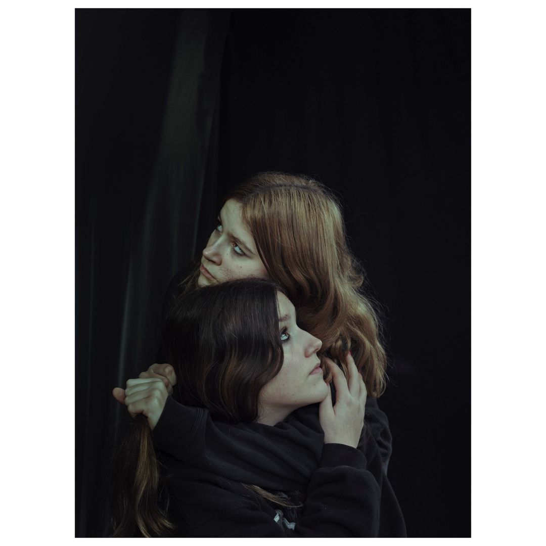 Two young women embrace in a photo studio setting. One woman has light auburn hair, the other dark brown. Both are looking upward.