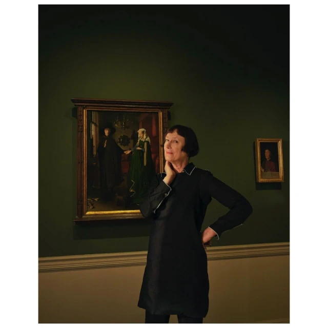 Mature woman in a black dress poses in front of framed paintings in a gallery with olive green walls and warm, indirect lighting.