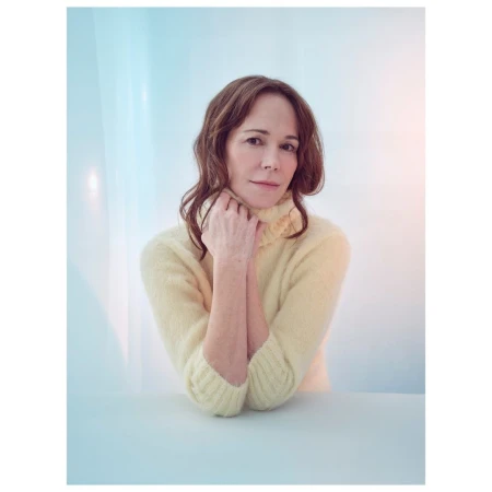 Woman in a yellow turtleneck sweater with hands held near her chin against a pale blue and white background