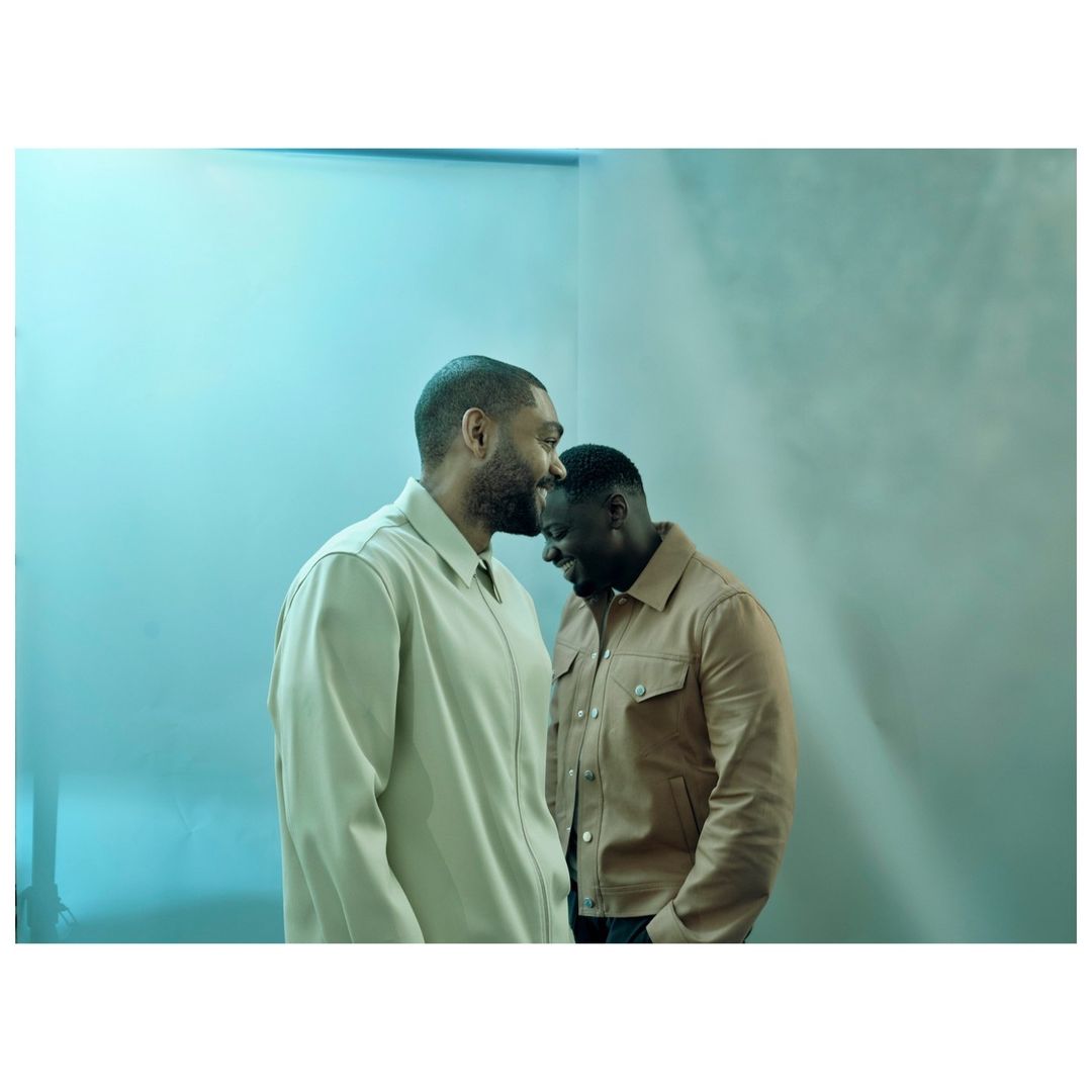 Two adult men in a semi-close shot indoors. The man on the left is wearing a light coloured jacket and smiling while the man on the right is wearing a tan jacket and is smiling.