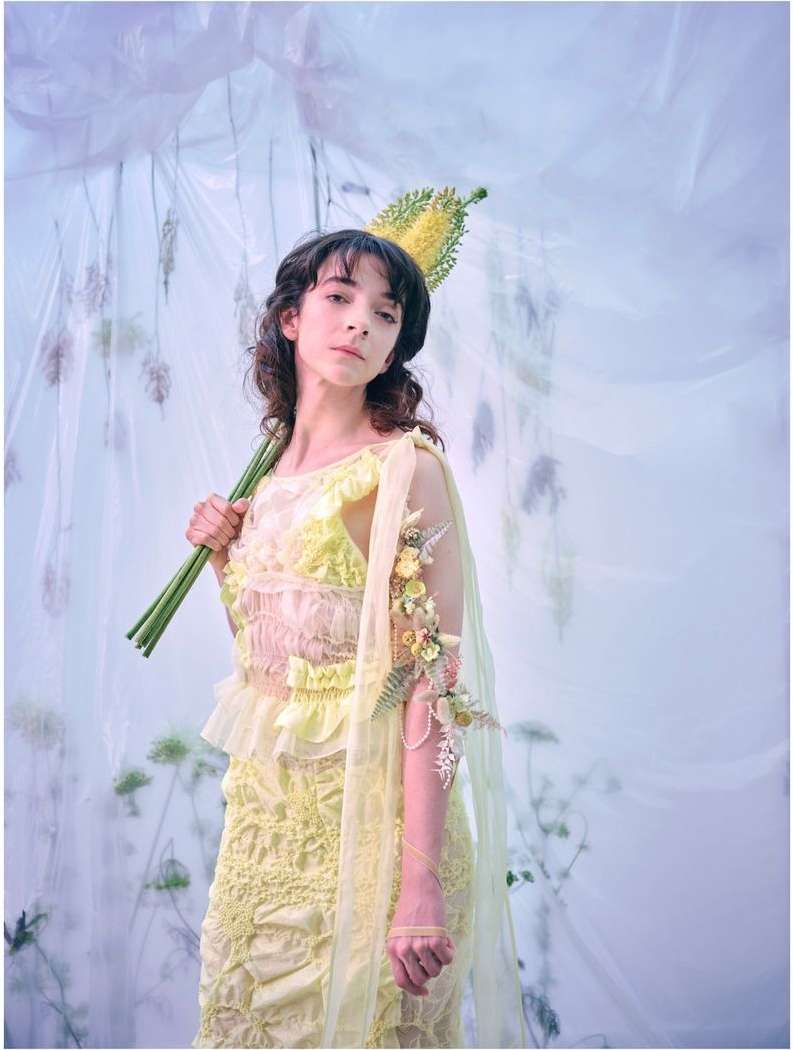 Portrait of a woman wearing a pale yellow dress and flower headdress, holding a floral staff, against a draped white background.